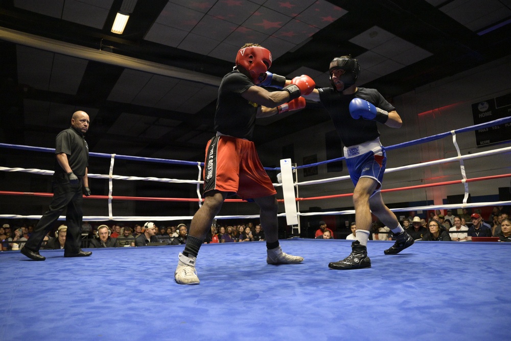 Two amateur boxers are engaged in an active exchange inside a boxing ring with blue canvas. One fighter wearing red gloves and red shorts throws a punch at an opponent in blue gloves and blue/white shorts, both wearing protective headgear. A referee in a black shirt monitors the bout in front of a large seated audience.