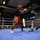 Two amateur boxers are engaged in an active exchange inside a boxing ring with blue canvas. One fighter wearing red gloves and red shorts throws a punch at an opponent in blue gloves and blue/white shorts, both wearing protective headgear. A referee in a black shirt monitors the bout in front of a large seated audience.