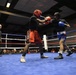 Two amateur boxers are engaged in an active exchange inside a boxing ring with blue canvas. One fighter wearing red gloves and red shorts throws a punch at an opponent in blue gloves and blue/white shorts, both wearing protective headgear. A referee in a black shirt monitors the bout in front of a large seated audience.
