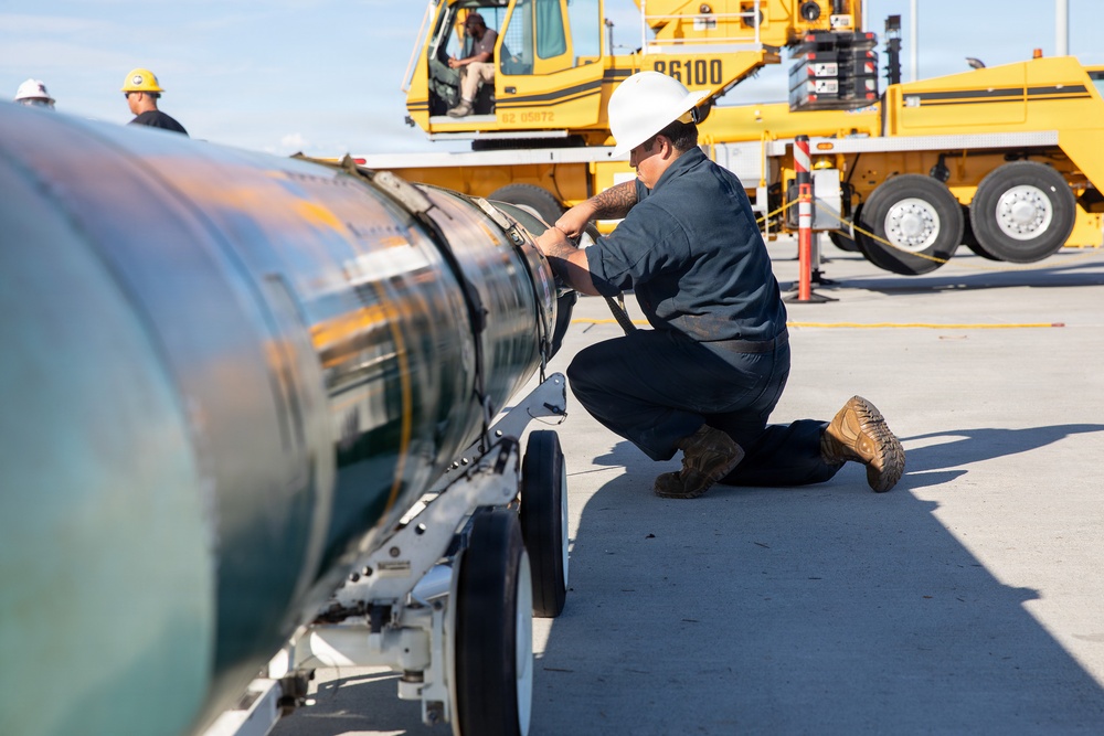 The image shows what appears to be military or aerospace ordnance/munitions handling on a tarmac or flight line. A technician in a white hard hat and dark coveralls is crouching beside a large cylindrical object on a wheeled cart, performing an inspection or maintenance task. A large yellow ground support vehicle numbered '86100' is visible in the background with additional personnel.