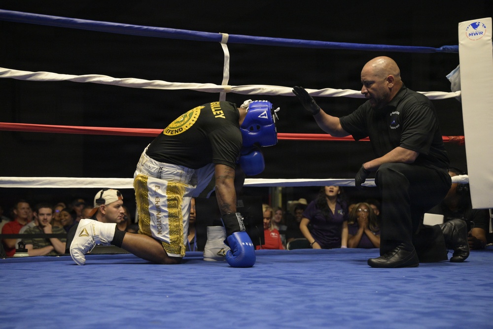 A boxing match is taking place in a ring where one fighter wearing a blue helmet and blue gloves is kneeling down, apparently knocked down or in distress. A referee in a black shirt is crouching nearby, appearing to check on the downed boxer or administer a count. The event appears to be an organized amateur boxing competition held in an indoor venue with a visible audience.