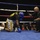 A boxing match is taking place in a ring where one fighter wearing a blue helmet and blue gloves is kneeling down, apparently knocked down or in distress. A referee in a black shirt is crouching nearby, appearing to check on the downed boxer or administer a count. The event appears to be an organized amateur boxing competition held in an indoor venue with a visible audience.