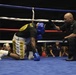 A boxing match is taking place in a ring where one fighter wearing a blue helmet and blue gloves is kneeling down, apparently knocked down or in distress. A referee in a black shirt is crouching nearby, appearing to check on the downed boxer or administer a count. The event appears to be an organized amateur boxing competition held in an indoor venue with a visible audience.