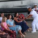 A U.S. Navy officer in dress white uniform is presenting a bouquet of red and pink roses to a seated woman in a navy blue dress during what appears to be a military ceremony. A second Navy officer in dress whites stands behind holding additional flowers. Several women and children are seated in folding chairs, likely family members attending the formal event.