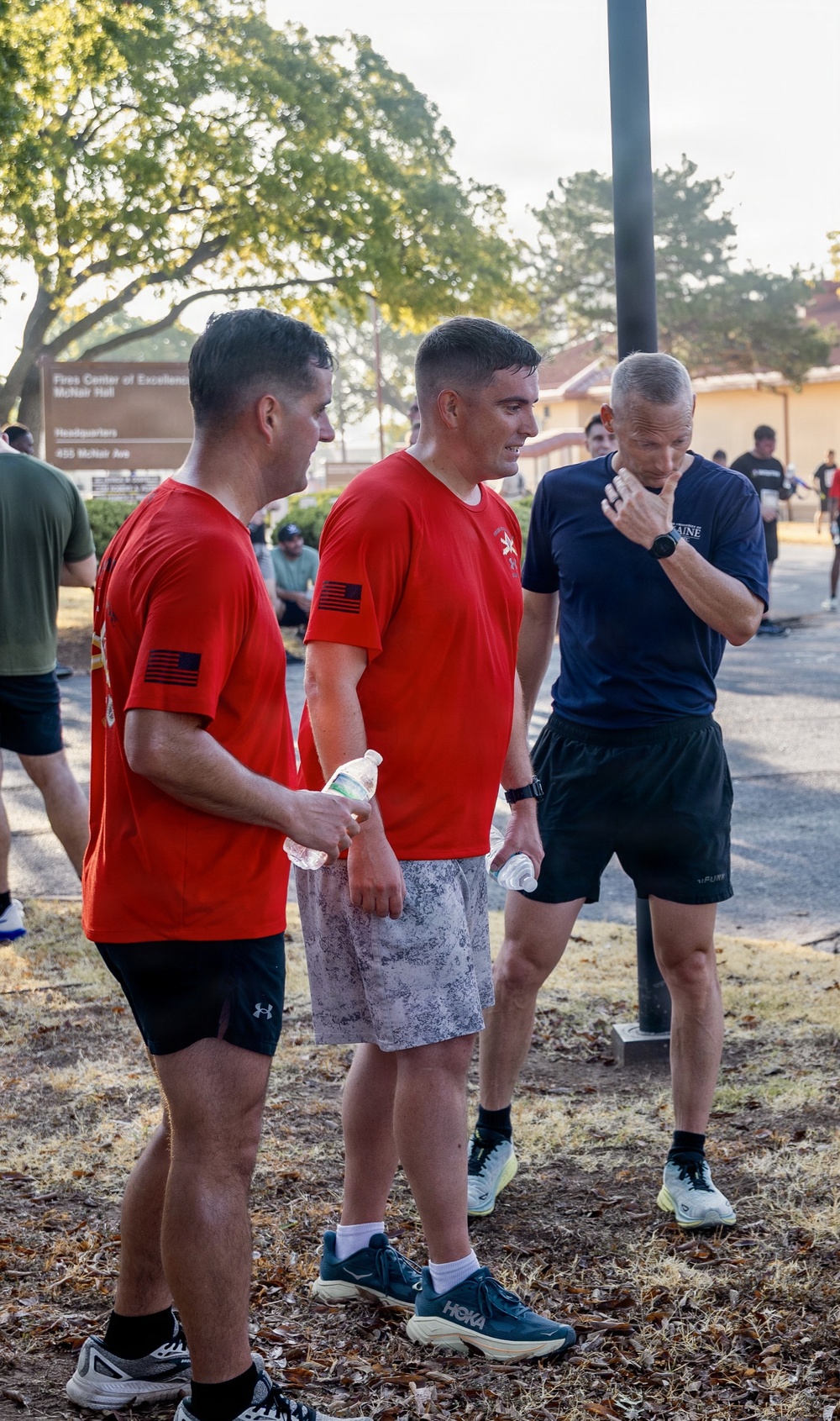 Three men in athletic wear are gathered outdoors on what appears to be a military installation, visible from the 'Fires Center of Excellence, McNair Hall, Headquarters' sign in the background. Two men are wearing matching red t-shirts with subdued American flag patches, suggesting a unit or team affiliation, while a third wears a navy blue shirt. The setting and attire suggest a post-run or physical training event.