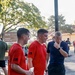 Three men in athletic wear are gathered outdoors on what appears to be a military installation, visible from the 'Fires Center of Excellence, McNair Hall, Headquarters' sign in the background. Two men are wearing matching red t-shirts with subdued American flag patches, suggesting a unit or team affiliation, while a third wears a navy blue shirt. The setting and attire suggest a post-run or physical training event.