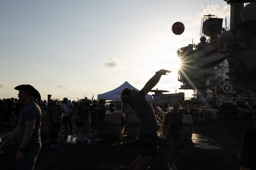 A group of people are gathered on what appears to be the flight deck of a naval aircraft carrier during a recreational or morale event. A person is captured mid-throw of a basketball, silhouetted against a dramatic sunburst near the ship's island superstructure. The scene suggests an organized crew recreation day or port visit event.