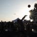 A group of people are gathered on what appears to be the flight deck of a naval aircraft carrier during a recreational or morale event. A person is captured mid-throw of a basketball, silhouetted against a dramatic sunburst near the ship's island superstructure. The scene suggests an organized crew recreation day or port visit event.