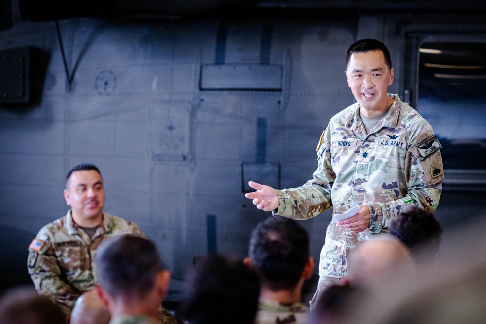A U.S. Army officer, identified by name tape as 'ROOKS,' is standing and addressing a group of soldiers in what appears to be a military briefing or informal talk inside a large military vehicle or aircraft hangar. The officer is smiling and gesturing with his right hand, holding what appears to be note cards. A second soldier in the background is seated and listening attentively.