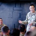 A U.S. Army officer, identified by name tape as 'ROOKS,' is standing and addressing a group of soldiers in what appears to be a military briefing or informal talk inside a large military vehicle or aircraft hangar. The officer is smiling and gesturing with his right hand, holding what appears to be note cards. A second soldier in the background is seated and listening attentively.