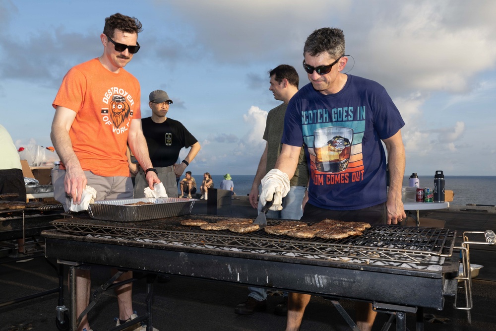 Two men are grilling meat on a large outdoor barbecue grill, appearing to be on the deck of a naval vessel or elevated outdoor platform near the ocean. The man on the left wears an orange 'Destroyer of Worlds' t-shirt, while the man on the right wears a navy blue 'Scotch Goes In, Wisdom Comes Out' t-shirt. Both are wearing protective oven mitts and are actively tending to multiple pieces of meat on the grill.