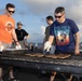 Two men are grilling meat on a large outdoor barbecue grill, appearing to be on the deck of a naval vessel or elevated outdoor platform near the ocean. The man on the left wears an orange 'Destroyer of Worlds' t-shirt, while the man on the right wears a navy blue 'Scotch Goes In, Wisdom Comes Out' t-shirt. Both are wearing protective oven mitts and are actively tending to multiple pieces of meat on the grill.