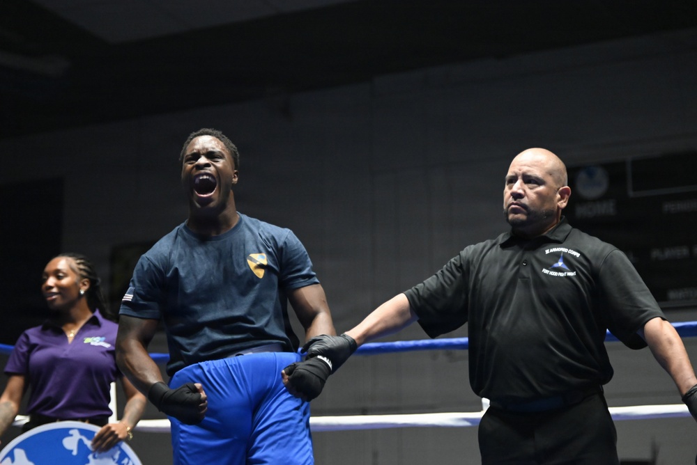 A boxer in a blue t-shirt and blue shorts celebrates with mouth open in a victory shout inside a boxing ring. A referee in a black polo shirt raises the boxer's hand to declare him the winner. A woman in a purple polo shirt stands in the background near the ring corner.