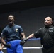 A boxer in a blue t-shirt and blue shorts celebrates with mouth open in a victory shout inside a boxing ring. A referee in a black polo shirt raises the boxer's hand to declare him the winner. A woman in a purple polo shirt stands in the background near the ring corner.