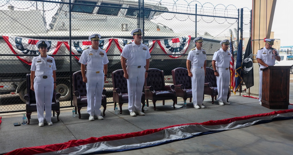 A formal military ceremony is taking place at what appears to be a naval facility or Coast Guard station. Six uniformed personnel in white dress uniforms are standing at attention on a raised platform, with one officer speaking at a podium. The setting is decorated with red, white, and blue patriotic bunting, and a naval vessel is visible in the background behind a chain-link fence.