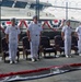 A formal military ceremony is taking place at what appears to be a naval facility or Coast Guard station. Six uniformed personnel in white dress uniforms are standing at attention on a raised platform, with one officer speaking at a podium. The setting is decorated with red, white, and blue patriotic bunting, and a naval vessel is visible in the background behind a chain-link fence.