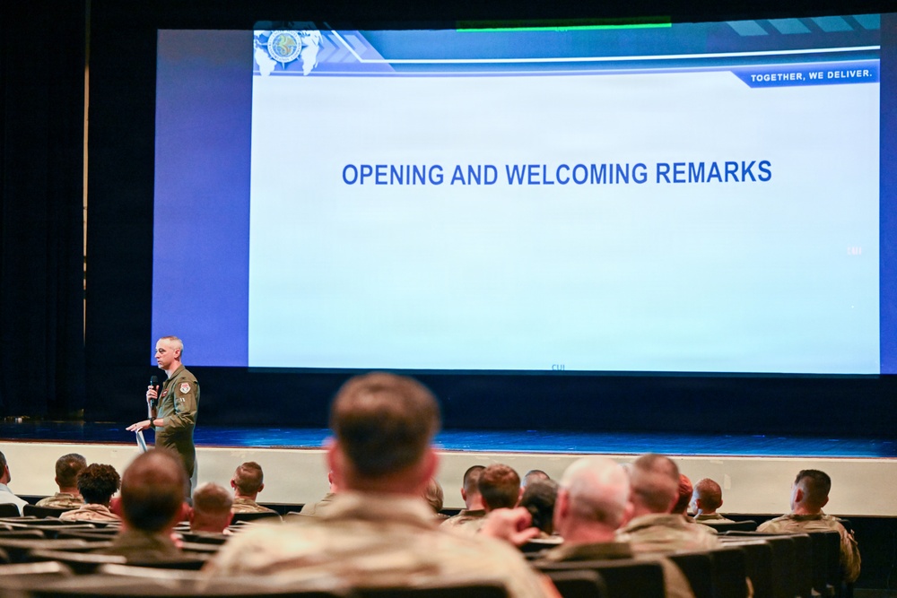 A military officer in olive/green flight suit is delivering opening and welcoming remarks at a formal military conference or briefing event. The speaker stands on a stage holding a microphone in front of a large projection screen displaying 'OPENING AND WELCOMING REMARKS' with a logo reading 'TOGETHER, WE DELIVER.' An audience of uniformed military personnel fills the auditorium seating.