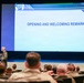 A military officer in olive/green flight suit is delivering opening and welcoming remarks at a formal military conference or briefing event. The speaker stands on a stage holding a microphone in front of a large projection screen displaying 'OPENING AND WELCOMING REMARKS' with a logo reading 'TOGETHER, WE DELIVER.' An audience of uniformed military personnel fills the auditorium seating.