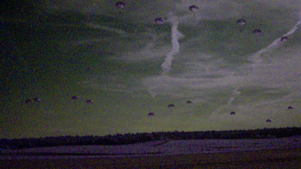 A low-light or night-vision style photograph showing a large-scale parachute drop over an open field or drop zone. Numerous parachutes are visible descending across the sky at various altitudes, suggesting a military airborne operation. The treeline is visible along the horizon, and the image has significant grain and color distortion consistent with low-light photography.