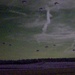 A low-light or night-vision style photograph showing a large-scale parachute drop over an open field or drop zone. Numerous parachutes are visible descending across the sky at various altitudes, suggesting a military airborne operation. The treeline is visible along the horizon, and the image has significant grain and color distortion consistent with low-light photography.