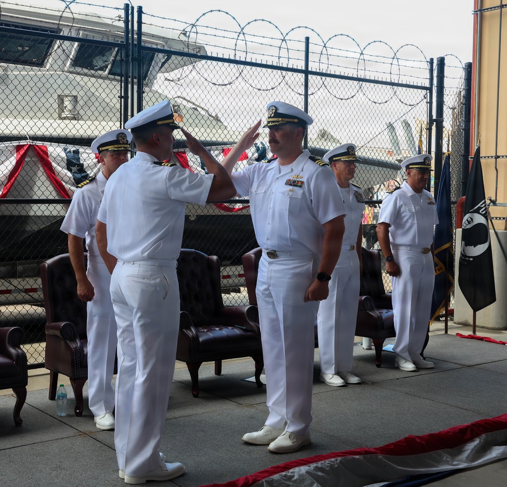 A formal U.S. Navy change of command or awards ceremony is taking place at what appears to be a naval installation or pier. Two officers in Service Dress White uniforms are exchanging salutes in the foreground, while additional personnel stand at attention in the background. A POW/MIA flag and other ceremonial elements are visible, indicating an official military ceremony.