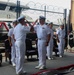 A formal U.S. Navy change of command or awards ceremony is taking place at what appears to be a naval installation or pier. Two officers in Service Dress White uniforms are exchanging salutes in the foreground, while additional personnel stand at attention in the background. A POW/MIA flag and other ceremonial elements are visible, indicating an official military ceremony.