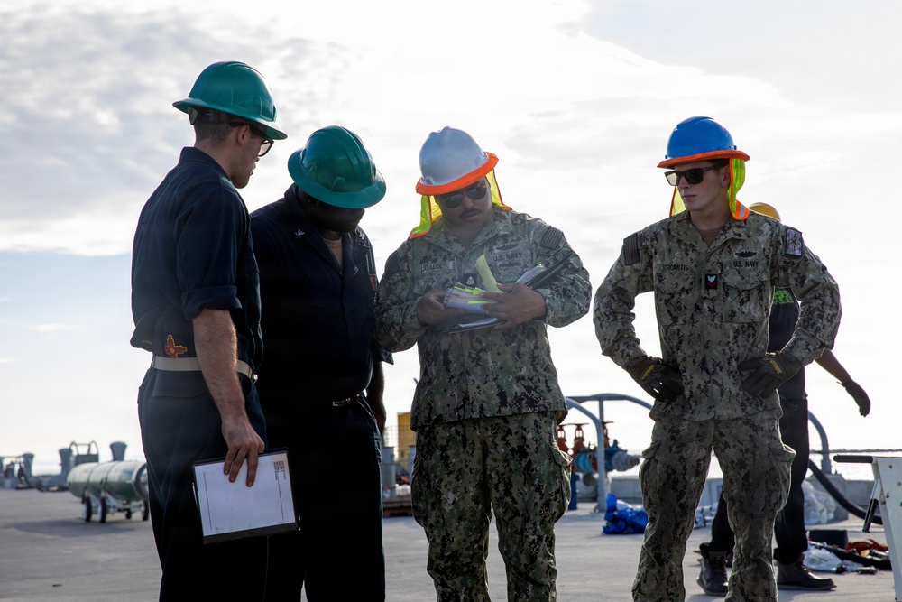 Four U.S. Navy personnel are gathered on what appears to be the flight deck of a naval vessel, reviewing documents or a clipboard. Two individuals are wearing camouflage NWU (Navy Working Uniform) while others wear dark navy uniforms, all equipped with hard hats and high-visibility safety gear. The setting suggests an operational briefing or inspection activity aboard a ship.
