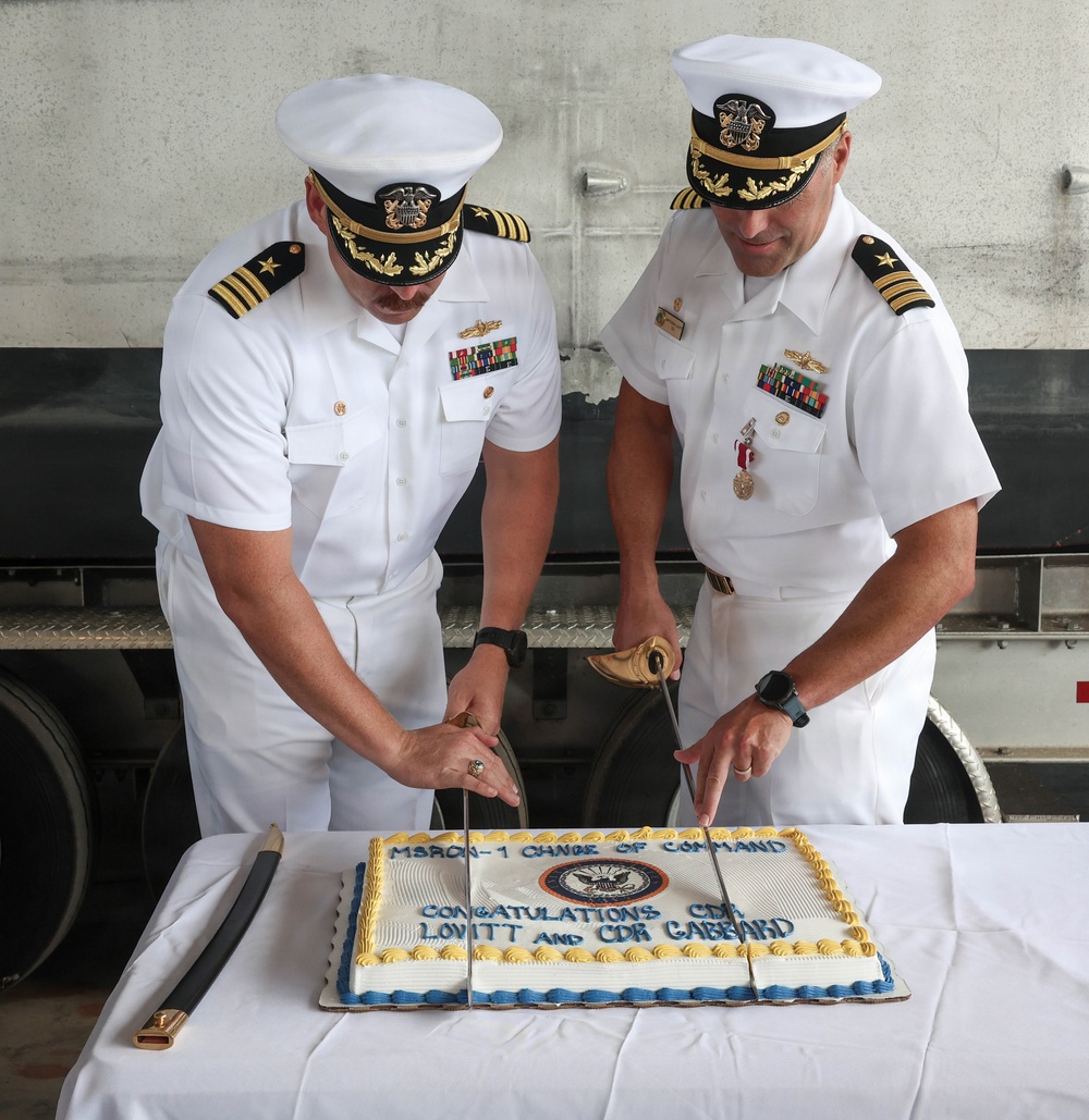 Two U.S. Navy Commanders in white dress uniforms are cutting a ceremonial cake with swords at a Change of Command ceremony for MSRON-1. The cake bears the text 'MSRON-1 CHANGE OF COMMAND' and 'CONGRATULATIONS CDR LOVITT AND CDR GABBARD,' along with a U.S. Navy emblem. Both officers display rank insignia, ribbons, and warfare qualification badges consistent with Commander (O-5) grade.