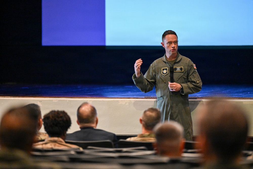 A military officer in an olive green flight suit is delivering a presentation or briefing to an audience in what appears to be an auditorium or theater setting. The speaker is holding a microphone and gesturing with one hand, standing in front of a large projection screen. The audience, visible from behind, consists of multiple individuals in various military and civilian attire.
