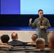 A military officer in an olive green flight suit is delivering a presentation or briefing to an audience in what appears to be an auditorium or theater setting. The speaker is holding a microphone and gesturing with one hand, standing in front of a large projection screen. The audience, visible from behind, consists of multiple individuals in various military and civilian attire.