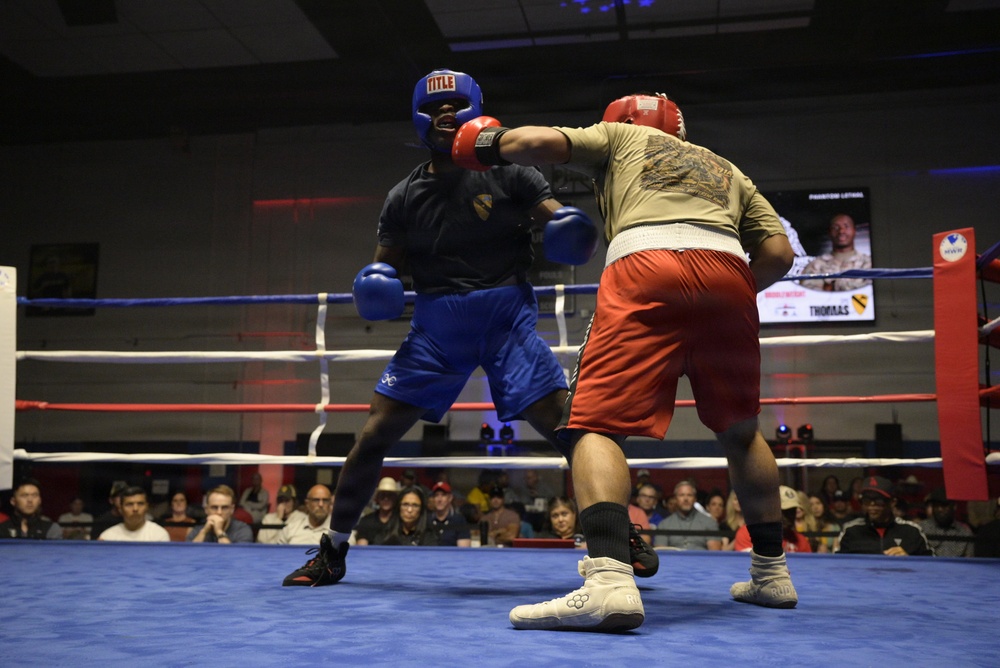 Two boxers are competing in an amateur boxing match inside an indoor ring with blue canvas and red/blue ropes. One boxer wears blue gear and shorts while the other wears red shorts and a tan shirt with red headgear. A crowd of spectators watches from the background, and a display screen is visible showing fighter information.