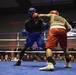 Two boxers are competing in an amateur boxing match inside an indoor ring with blue canvas and red/blue ropes. One boxer wears blue gear and shorts while the other wears red shorts and a tan shirt with red headgear. A crowd of spectators watches from the background, and a display screen is visible showing fighter information.