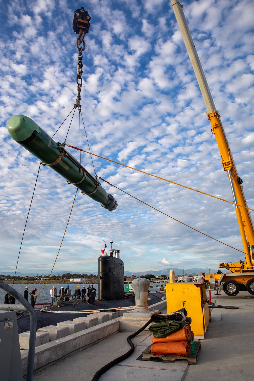 A torpedo is being loaded onto a submarine at a naval pier using a large yellow mobile crane. Personnel in safety gear are visible on the submarine's deck and along the pier, overseeing the weapons loading operation. The setting appears to be a U.S. Navy installation, likely on the West Coast based on the visible mountain range in the background.