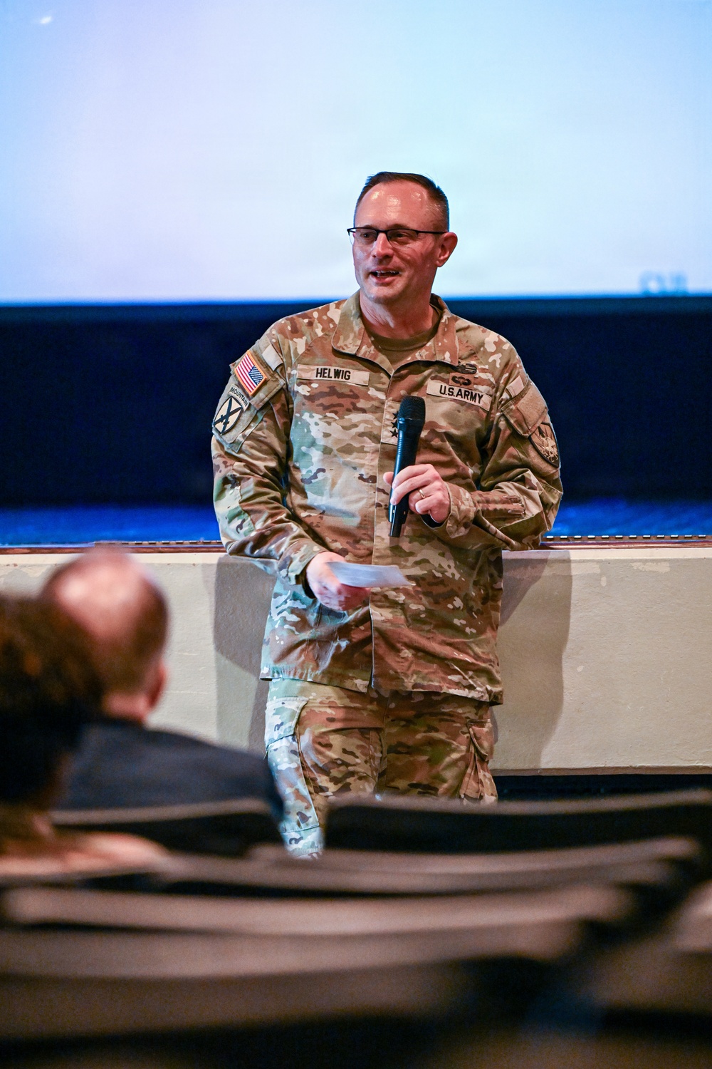A U.S. Army soldier named Helwig, wearing Operational Camouflage Pattern (OCP) uniform, is speaking into a handheld microphone at what appears to be a formal briefing or presentation event. He is holding papers in one hand and a microphone in the other, standing in front of a projection screen. An audience member is visible in the foreground, seated with their back to the camera.