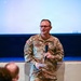 A U.S. Army soldier named Helwig, wearing Operational Camouflage Pattern (OCP) uniform, is speaking into a handheld microphone at what appears to be a formal briefing or presentation event. He is holding papers in one hand and a microphone in the other, standing in front of a projection screen. An audience member is visible in the foreground, seated with their back to the camera.