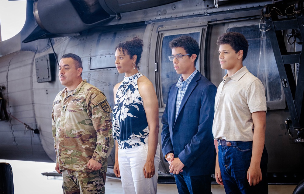 Four individuals stand in front of a military helicopter, likely inside a hangar. One person on the left is wearing U.S. Army camouflage (OCP/MultiCam) uniform, while the other three are dressed in civilian attire. The setting and posture suggest a formal military ceremony or family recognition event.