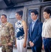 Four individuals stand in front of a military helicopter, likely inside a hangar. One person on the left is wearing U.S. Army camouflage (OCP/MultiCam) uniform, while the other three are dressed in civilian attire. The setting and posture suggest a formal military ceremony or family recognition event.