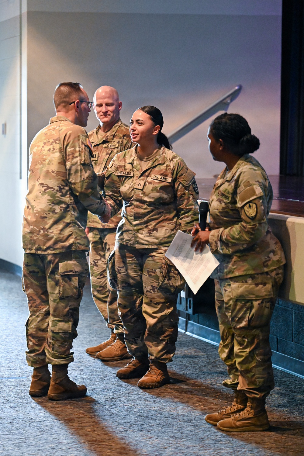 Four U.S. Army soldiers in OCP (Operational Camouflage Pattern) uniforms are gathered in what appears to be an auditorium or meeting facility. A female soldier with the name tape 'GONZALES' and a Private First Class rank insignia is shaking hands with another soldier, suggesting a recognition or award ceremony. A fourth soldier to the right holds a microphone and papers, indicating a formal event with a presenter or emcee.