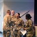 Four U.S. Army soldiers in OCP (Operational Camouflage Pattern) uniforms are gathered in what appears to be an auditorium or meeting facility. A female soldier with the name tape 'GONZALES' and a Private First Class rank insignia is shaking hands with another soldier, suggesting a recognition or award ceremony. A fourth soldier to the right holds a microphone and papers, indicating a formal event with a presenter or emcee.