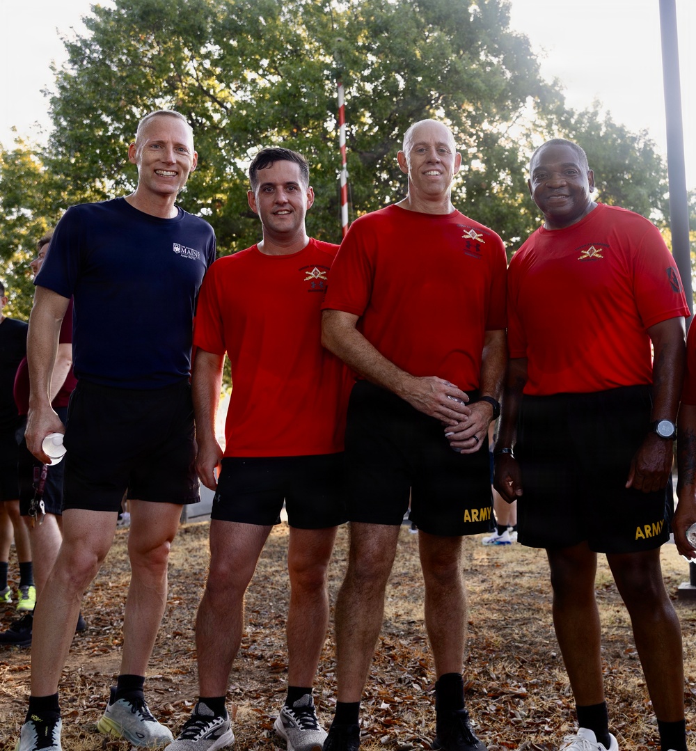 Four men pose together outdoors in athletic/PT (physical training) attire. Three are wearing matching red shirts with what appears to be a military insignia, and two are wearing shorts with 'ARMY' printed on them, suggesting a U.S. Army physical training event. The setting appears to be an outdoor area with trees and fallen leaves in the background.