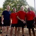 Four men pose together outdoors in athletic/PT (physical training) attire. Three are wearing matching red shirts with what appears to be a military insignia, and two are wearing shorts with 'ARMY' printed on them, suggesting a U.S. Army physical training event. The setting appears to be an outdoor area with trees and fallen leaves in the background.