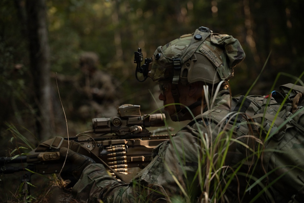 A soldier in MultiCam camouflage is positioned in a prone or low-crouch stance in a wooded environment, manning what appears to be a belt-fed machine gun. The soldier is equipped with advanced combat gear including a helmet-mounted device, optics on the weapon, and full tactical kit. A second soldier is visible in the blurred background, suggesting a small unit tactical exercise or field training operation.