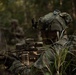 A soldier in MultiCam camouflage is positioned in a prone or low-crouch stance in a wooded environment, manning what appears to be a belt-fed machine gun. The soldier is equipped with advanced combat gear including a helmet-mounted device, optics on the weapon, and full tactical kit. A second soldier is visible in the blurred background, suggesting a small unit tactical exercise or field training operation.