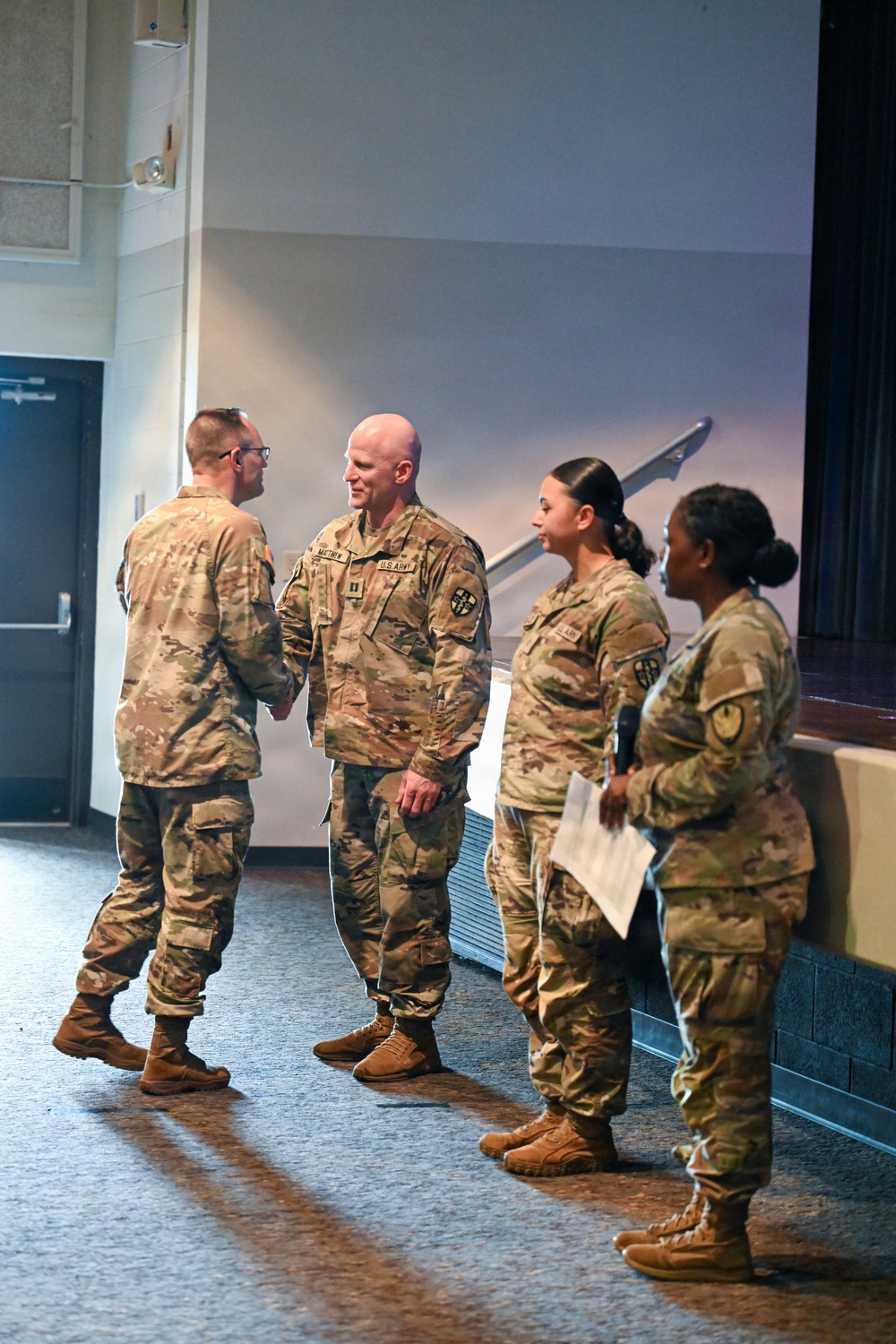 Four U.S. Army soldiers in Operational Camouflage Pattern (OCP) uniforms are gathered in what appears to be an auditorium or multipurpose room. Two soldiers on the left appear to be shaking hands or engaged in a formal greeting, while two female soldiers stand to the right, one holding papers and a microphone. The setting suggests a formal military ceremony or official event.