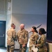Four U.S. Army soldiers in Operational Camouflage Pattern (OCP) uniforms are gathered in what appears to be an auditorium or multipurpose room. Two soldiers on the left appear to be shaking hands or engaged in a formal greeting, while two female soldiers stand to the right, one holding papers and a microphone. The setting suggests a formal military ceremony or official event.