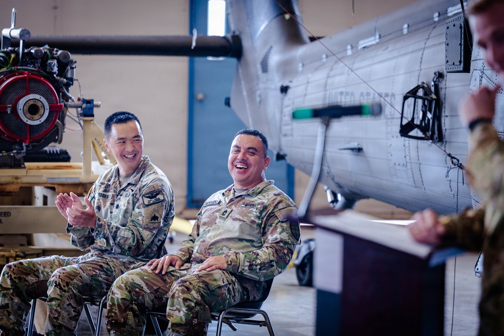Two U.S. Army soldiers in OCP (Operational Camouflage Pattern) uniforms are seated and laughing during what appears to be a military ceremony or briefing inside an aircraft hangar. A third uniformed individual stands at a podium to the right. The setting includes military aircraft components and equipment visible in the background.