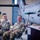 Sgt. Maj. Antonio Acevedo Promotion Ceremony Two U.S. Army soldiers in OCP (Operational Camouflage Pattern) uniforms are seated and laughing during what appears to be a military ceremony or briefing inside an aircraft hangar. A third uniformed individual stands at a podium to the right. The setting includes military aircraft components and equipment visible in the background.