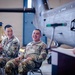 Two U.S. Army soldiers in OCP (Operational Camouflage Pattern) uniforms are seated and laughing during what appears to be a military ceremony or briefing inside an aircraft hangar. A third uniformed individual stands at a podium to the right. The setting includes military aircraft components and equipment visible in the background.