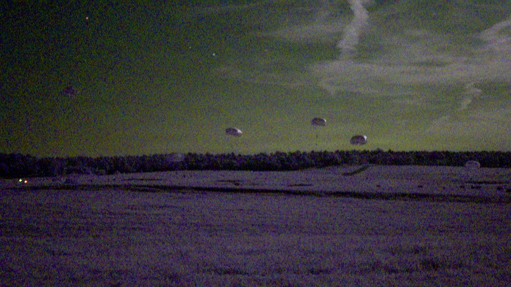 A low-light nighttime photograph showing what appears to be a military or training parachute drop over an open field. Multiple parachutes are visible descending near the horizon line against a dark, greenish-tinted sky. The image is heavily grainy and noisy, consistent with a high-ISO or night-vision-adjacent camera capture.