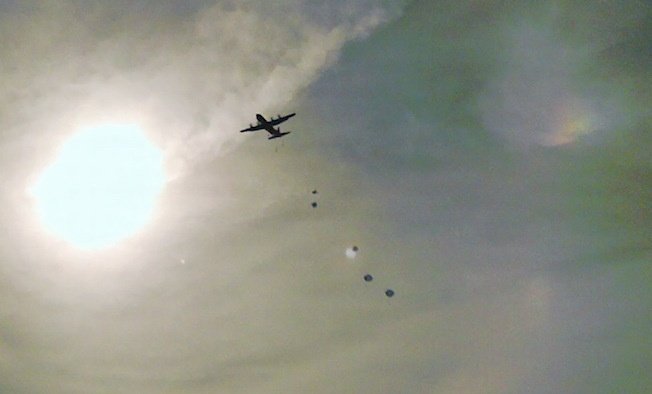 A large military transport aircraft, consistent with a C-130 Hercules silhouette, is photographed from below against a bright, hazy sky with the sun visible at left. The aircraft appears to be conducting an airdrop operation, with multiple parachutes or cargo bundles visible descending below the aircraft. The image is backlit and slightly overexposed due to the sun's proximity in the frame.